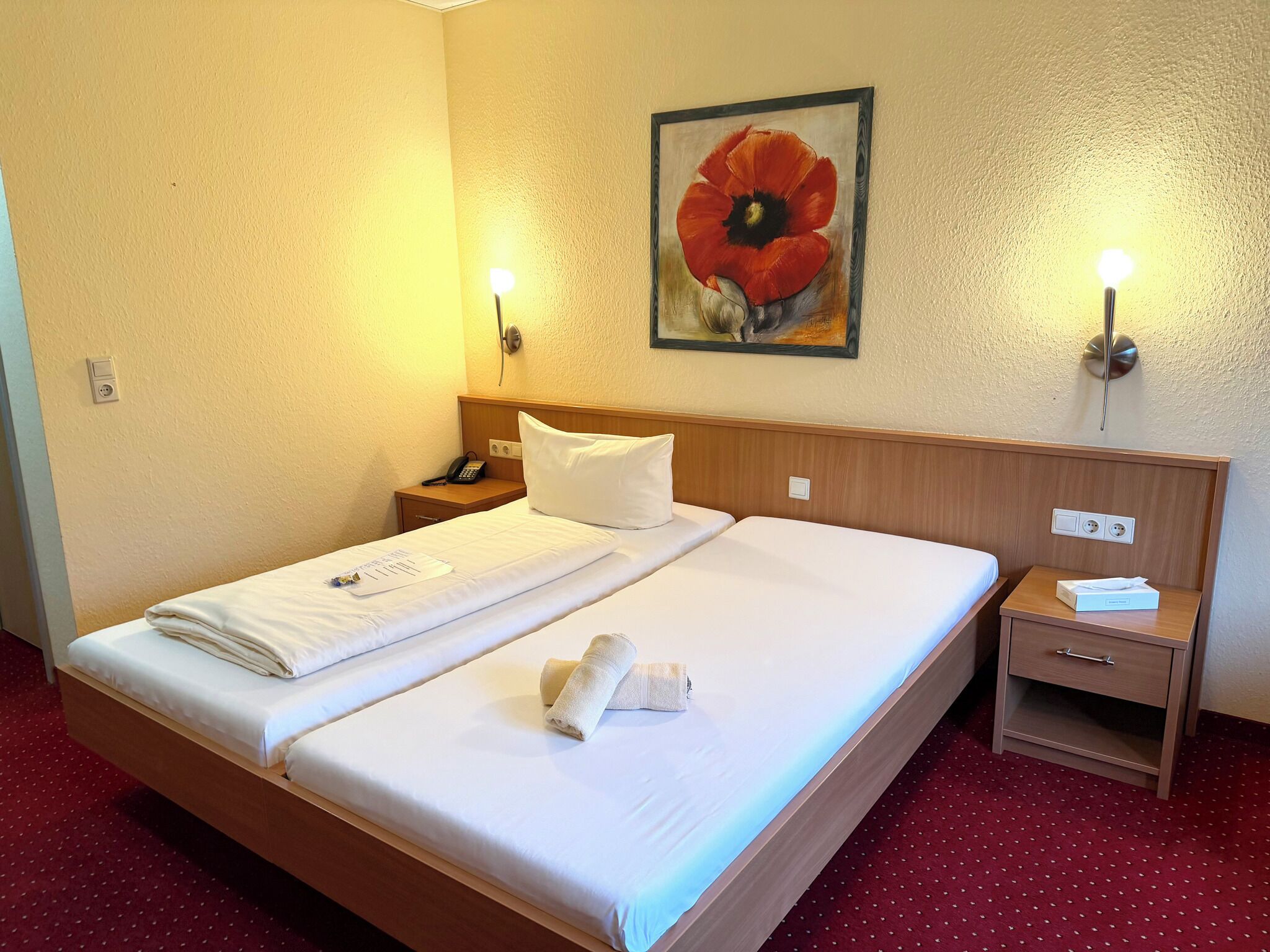 A hotel room with a large bed, a bedside table with a lamp, a painting on the wall, and a red carpet.