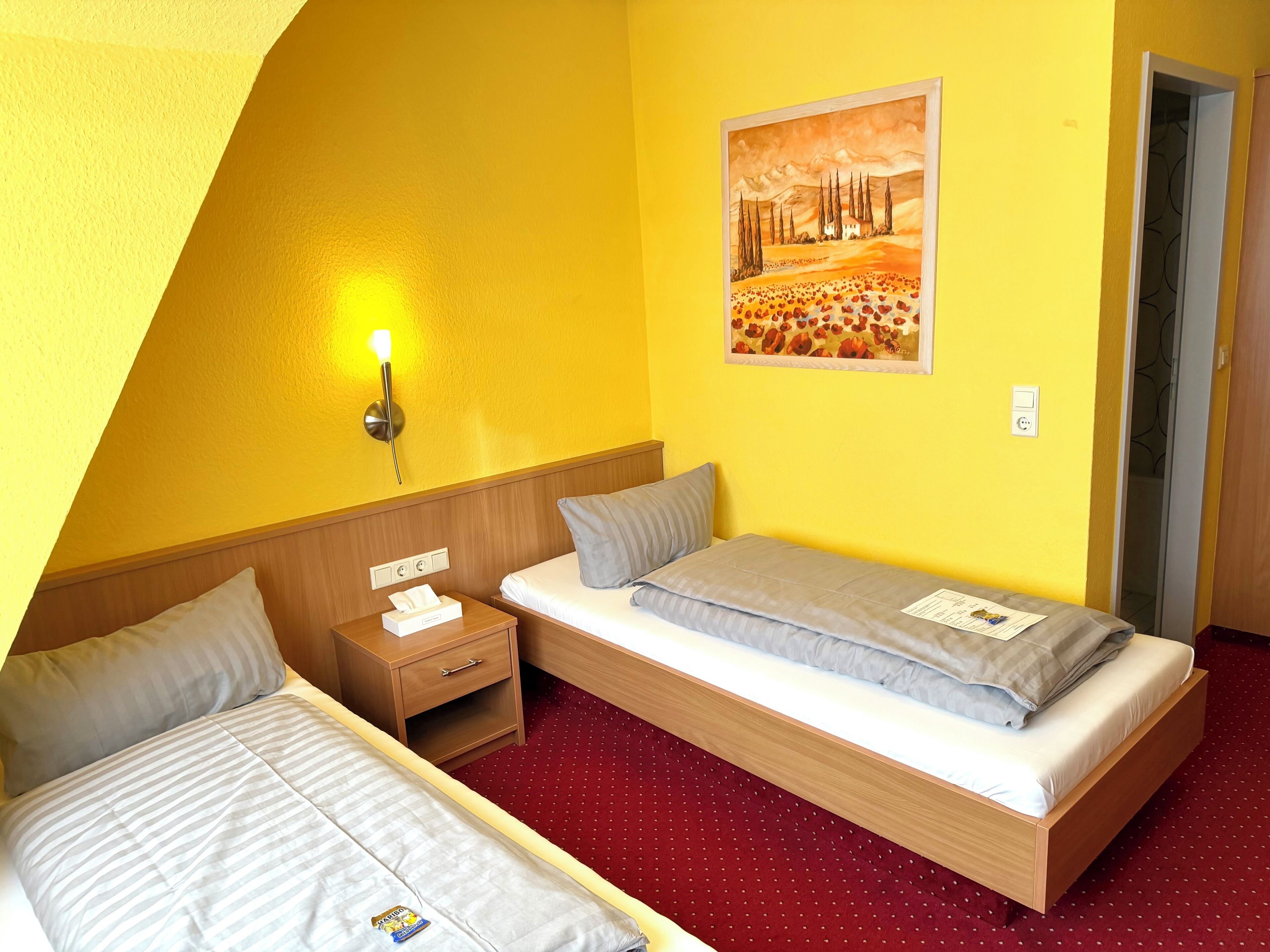 A hotel room with two single beds, a wooden nightstand with a lamp, a painting on the wall, and a red carpeted floor.
