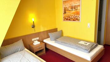 A hotel room with two single beds, a wooden nightstand with a lamp, a painting on the wall, and a red carpeted floor.