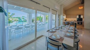 A modern dining area with a long table set for a meal, large windows offering a view of a pool and greenery, and a kitchen in the background. - Oceanfront 10BR Lux Villa • Pool • Full Ocean View (Noord)
