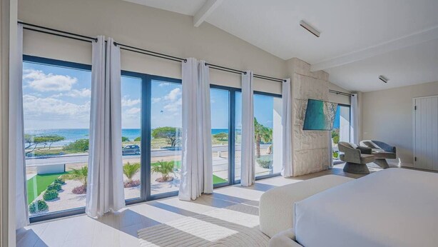 A modern bedroom with a large bed, a flat-screen TV mounted on the wall, and a view of the ocean through floor-to-ceiling windows. - Oceanfront 10BR Lux Villa • Pool • Full Ocean View (Noord)