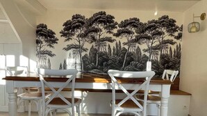 A dining area with a long table and white chairs, a mural of trees on the wall, and a light fixture on the wall. - Le Petit Shamrock - exceptional villa on the beach (Cabourg)
