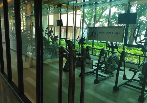 Une salle de sport équipée de divers appareils d’exercice, un panneau indiquant l’espace réservé aux entraînements, et une vue sur la verdure à l’extérieur.