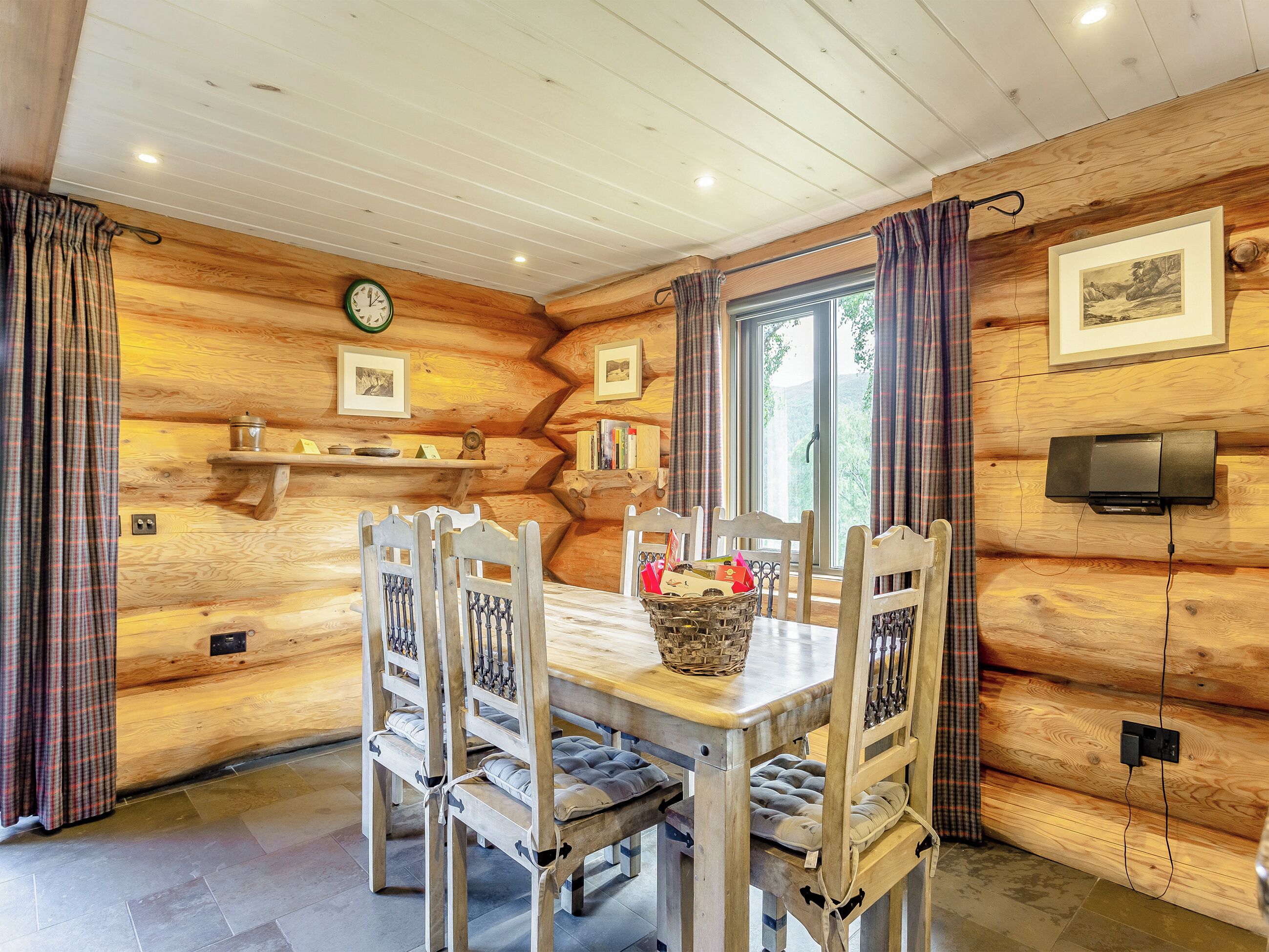 A wooden interior with a dining table, chairs, a clock, framed pictures, and a television.
