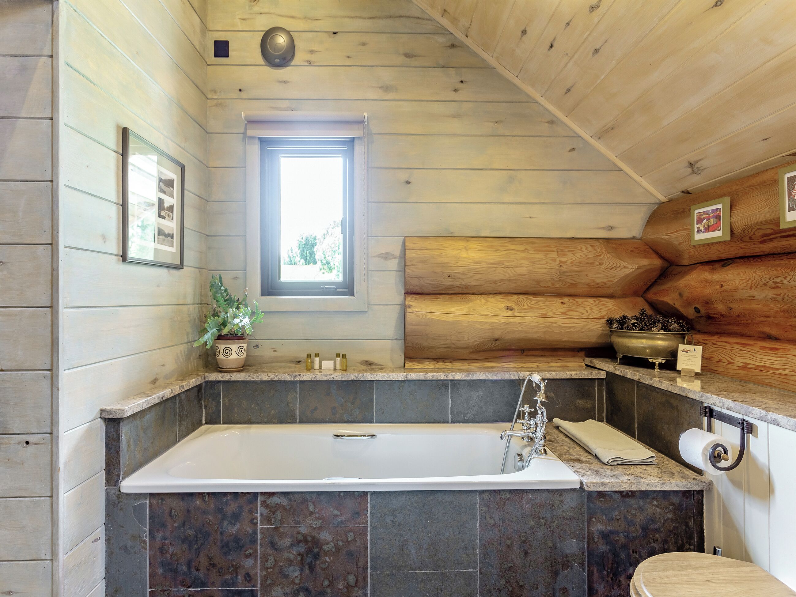 A log cabin bathroom with a bathtub, a window, a sink with toiletries, and a potted plant.