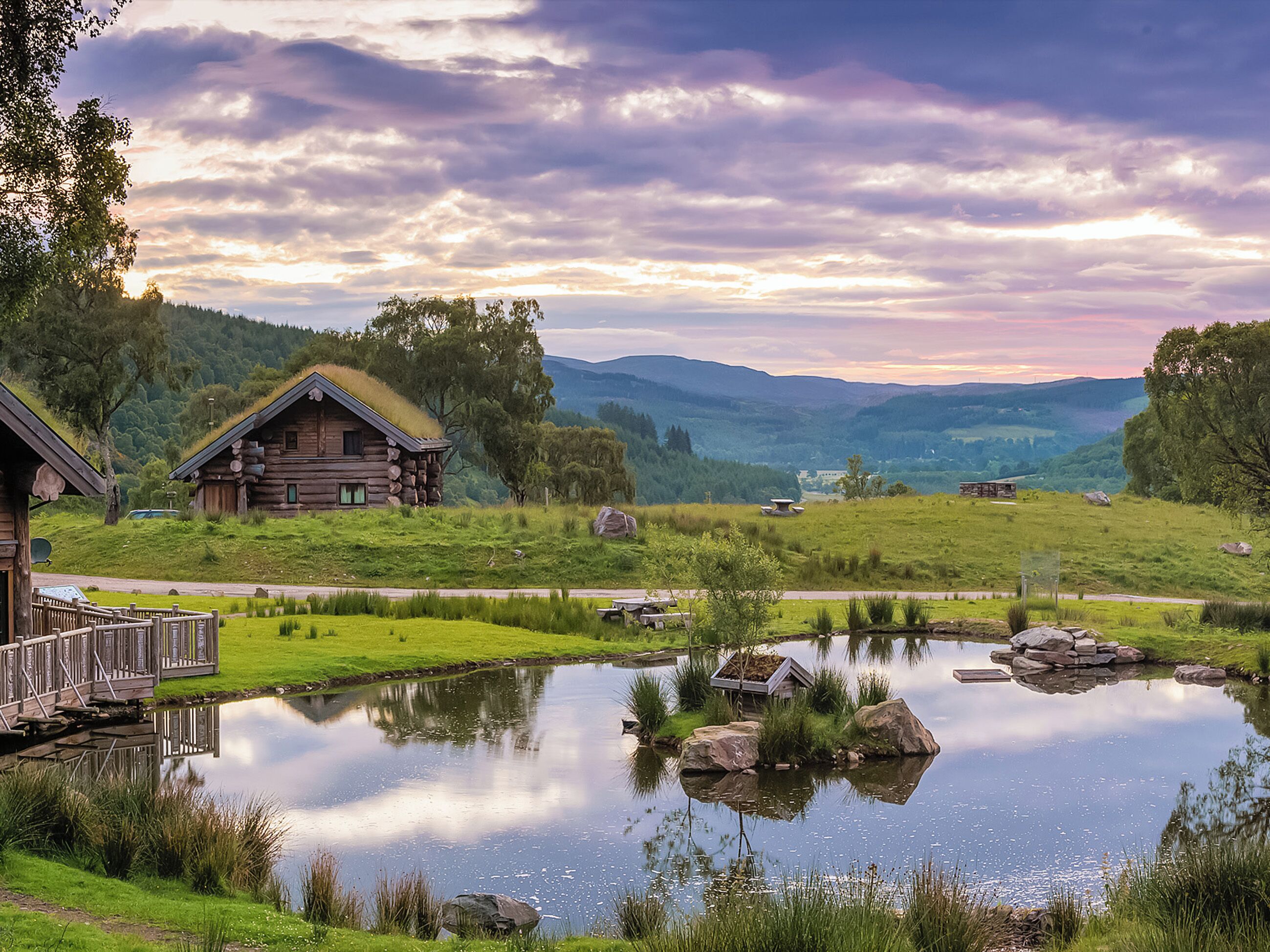 A log cabin with a deck, a pond with rocks, and a view of the surrounding hills.
