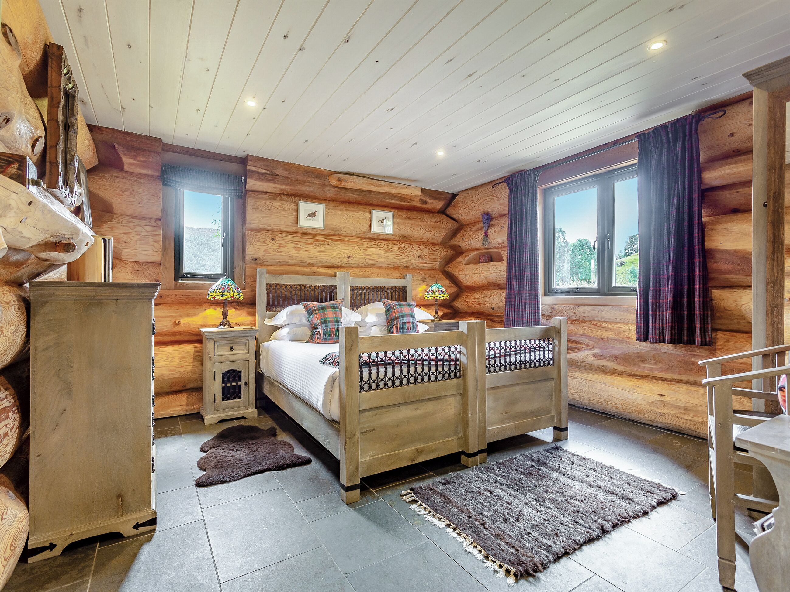 Un dormitorio tipo cabaña de madera con una cama de madera, mesitas de noche, un tapete y una ventana con cortinas.