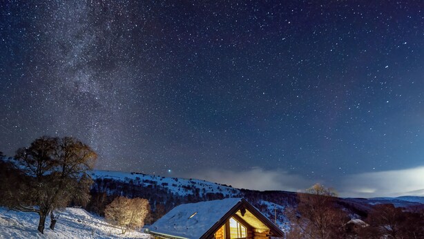 Una cabaña de troncos con un porche y un paisaje nevado bajo un cielo estrellado.