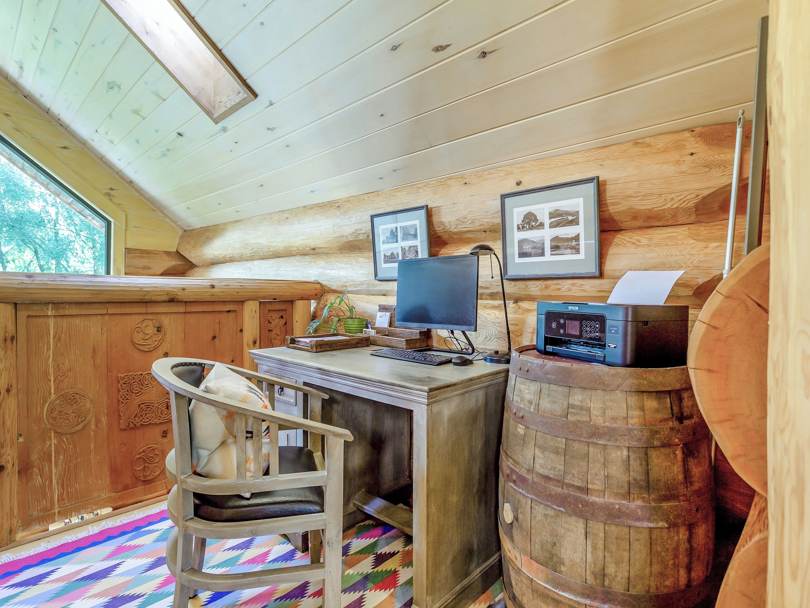 A rustic cabin interior with a wooden desk, computer, chair, and a barrel.