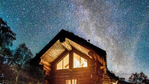 Una cabaña de troncos con ventanas amplias, una terraza de madera y un cielo nocturno estrellado.