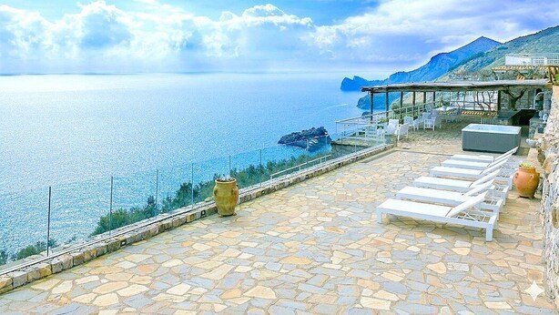 Une terrasse en bord de mer avec un sol en pierre, des chaises longues, une balustrade en verre et une vue sur la mer et les montagnes lointaines.