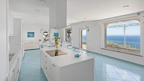 Une cuisine moderne dotée d’un grand îlot central, de meubles blancs et offrant une vue sur la mer grâce à une grande fenêtre.