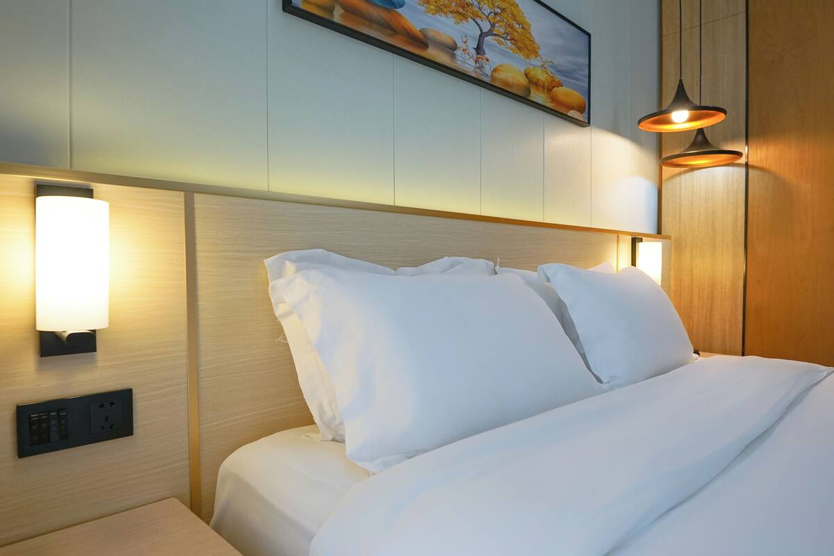 Una cama bien hecha con sábanas blancas, una lámpara de pared y un cuadro sobre la cama.