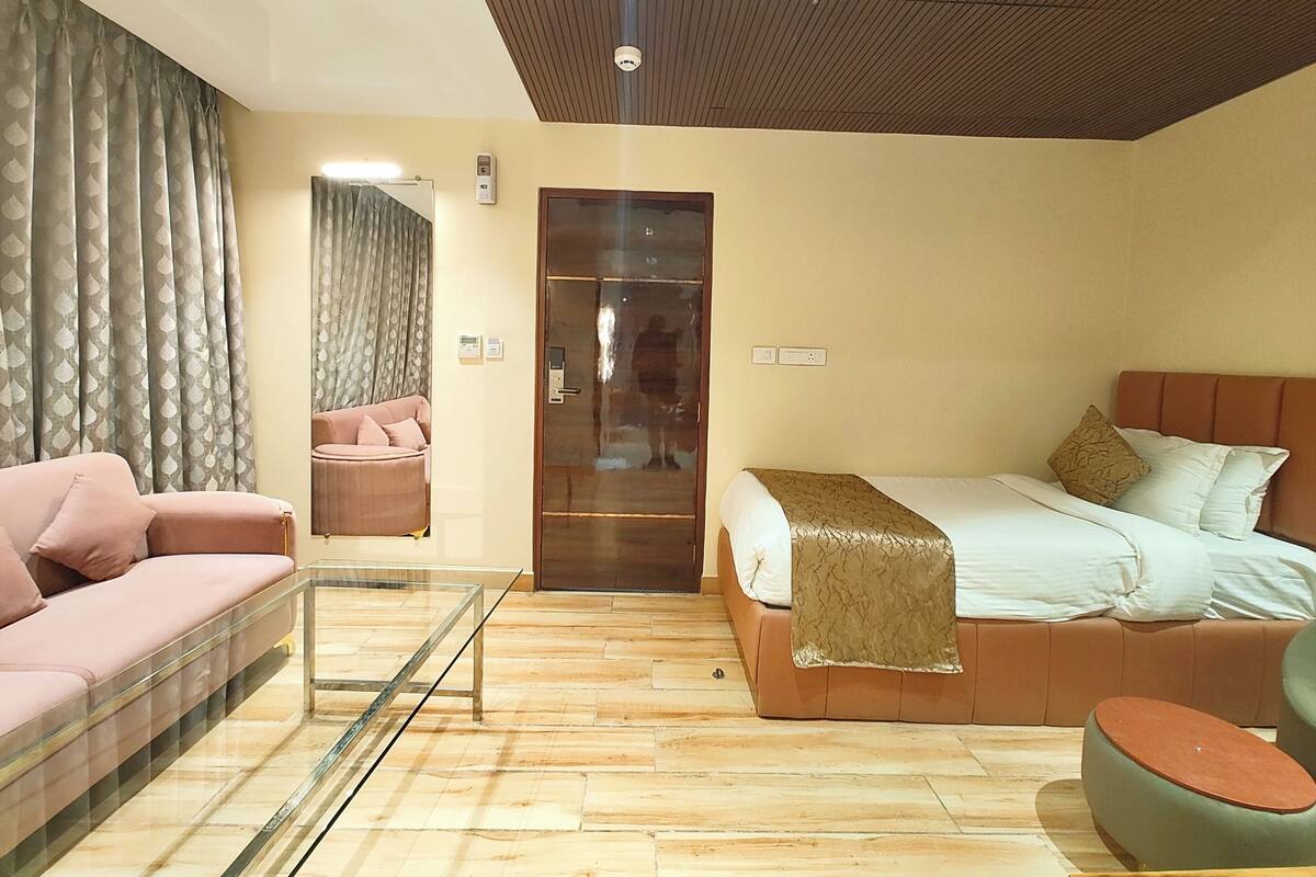 A modern hotel room with a glass coffee table, a pink sofa, a bed with a brown headboard, and a green ottoman.