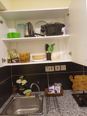 A kitchen with a sink, a stove, and a countertop. There are various kitchen items on the shelves, including a green basket, a black pot, and a wooden cutting board. - Cozy Studio near JKIA, SGR, Expressway /Mombasa Road, Syokimau. Luxore Apartment (Nairobi)