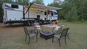 Un véhicule récréatif équipé d’un coin cuisine, d’un espace repas avec chaises et table, et d’un petit foyer extérieur.