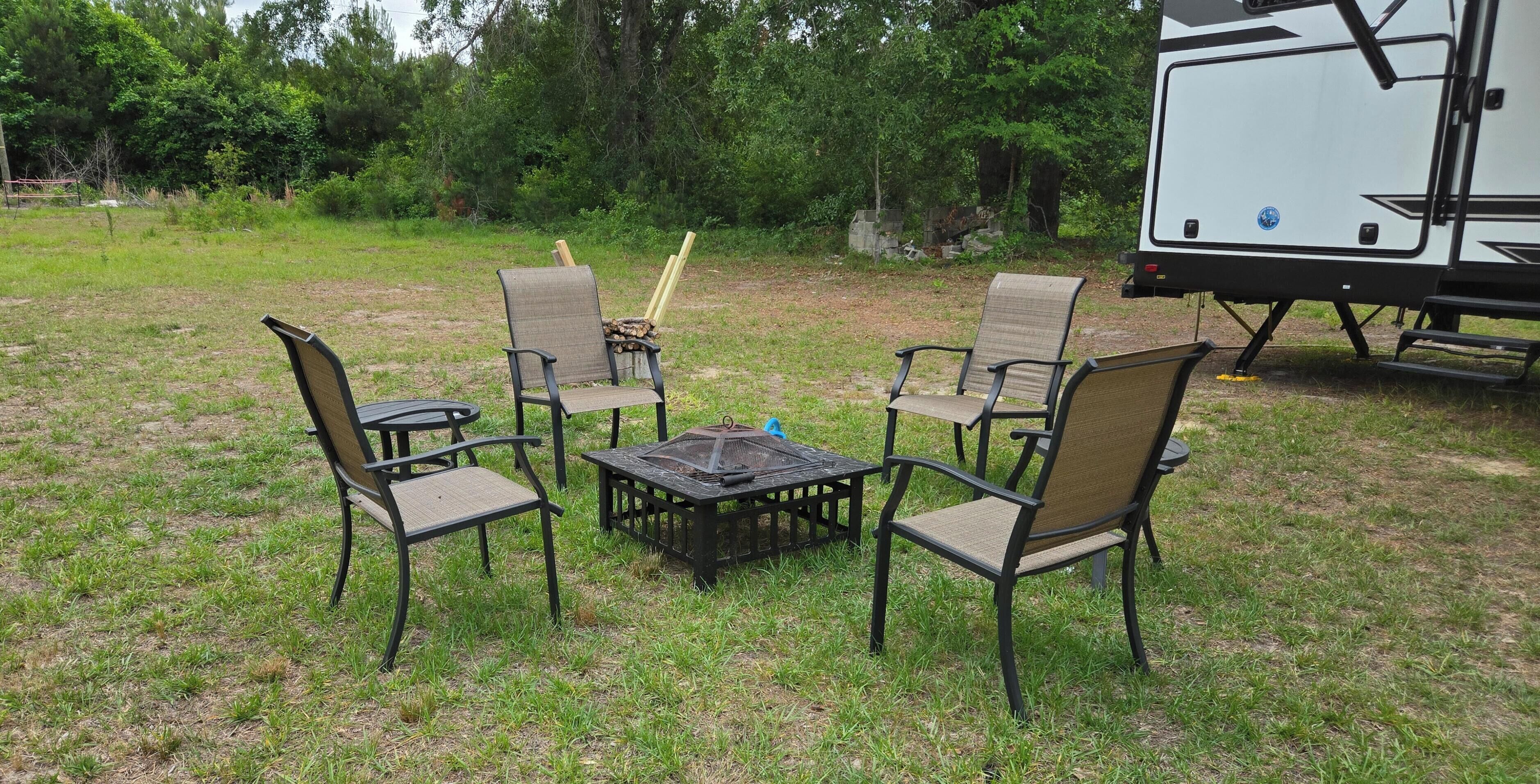 A campsite with four chairs and a table, surrounded by grass and trees, with a camper parked nearby.