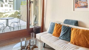Una sala de estar moderna con una mesa de centro de vidrio, un sofá blanco con cojines coloridos y un amplio ventanal que ofrece vistas a un patio y edificios.