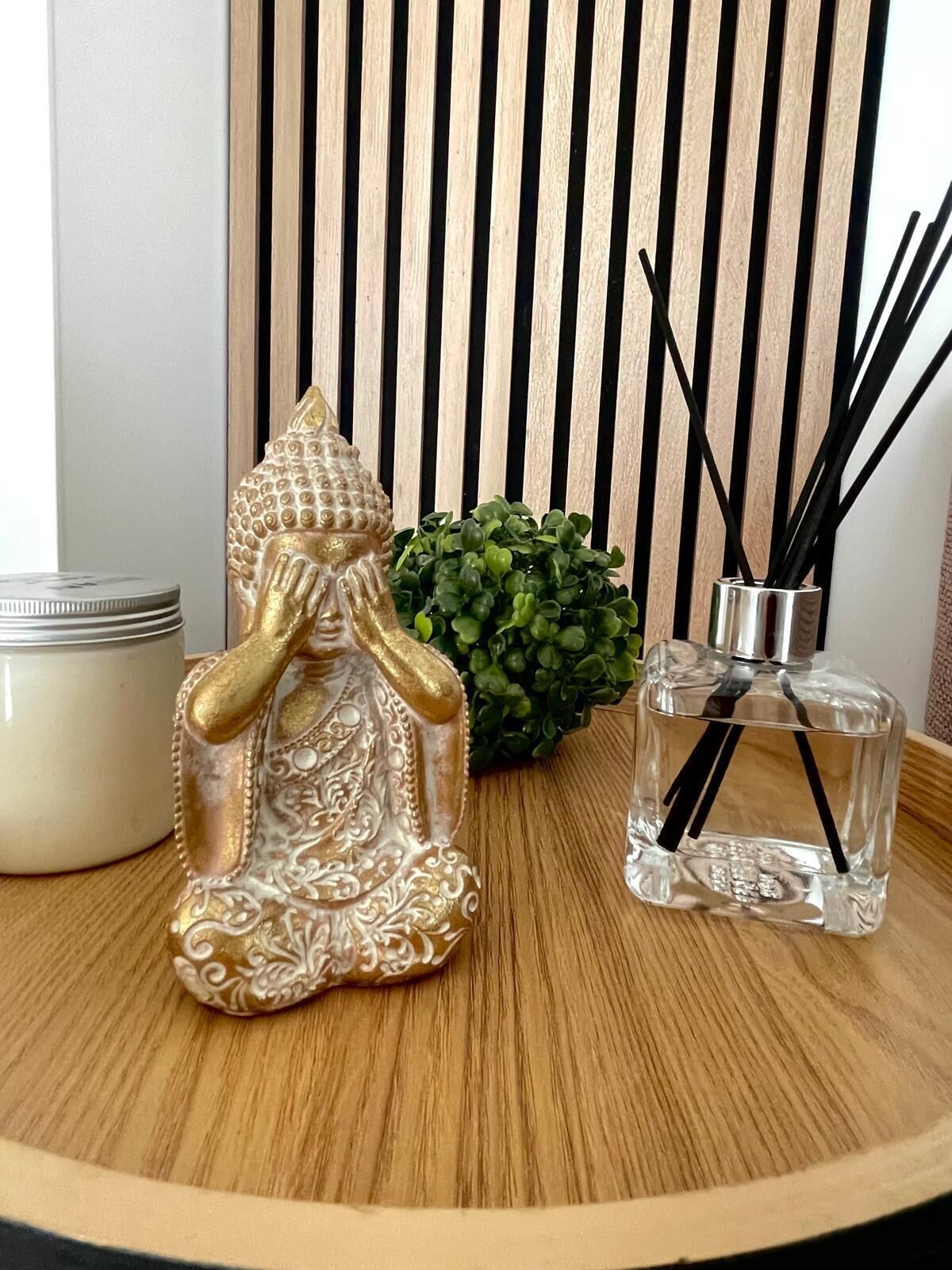 Une statue de Bouddha décorative, une petite plante en pot, une bougie et un flacon en verre contenant des tiges de roseau, le tout posé sur une surface en bois.