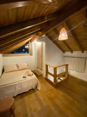 A bedroom with a sloped ceiling, wooden beams, a bed with beige bedding, a small wooden table, and a hanging light fixture. - Pitanga House - Ponta do Papagaio Beach (Palhoça)