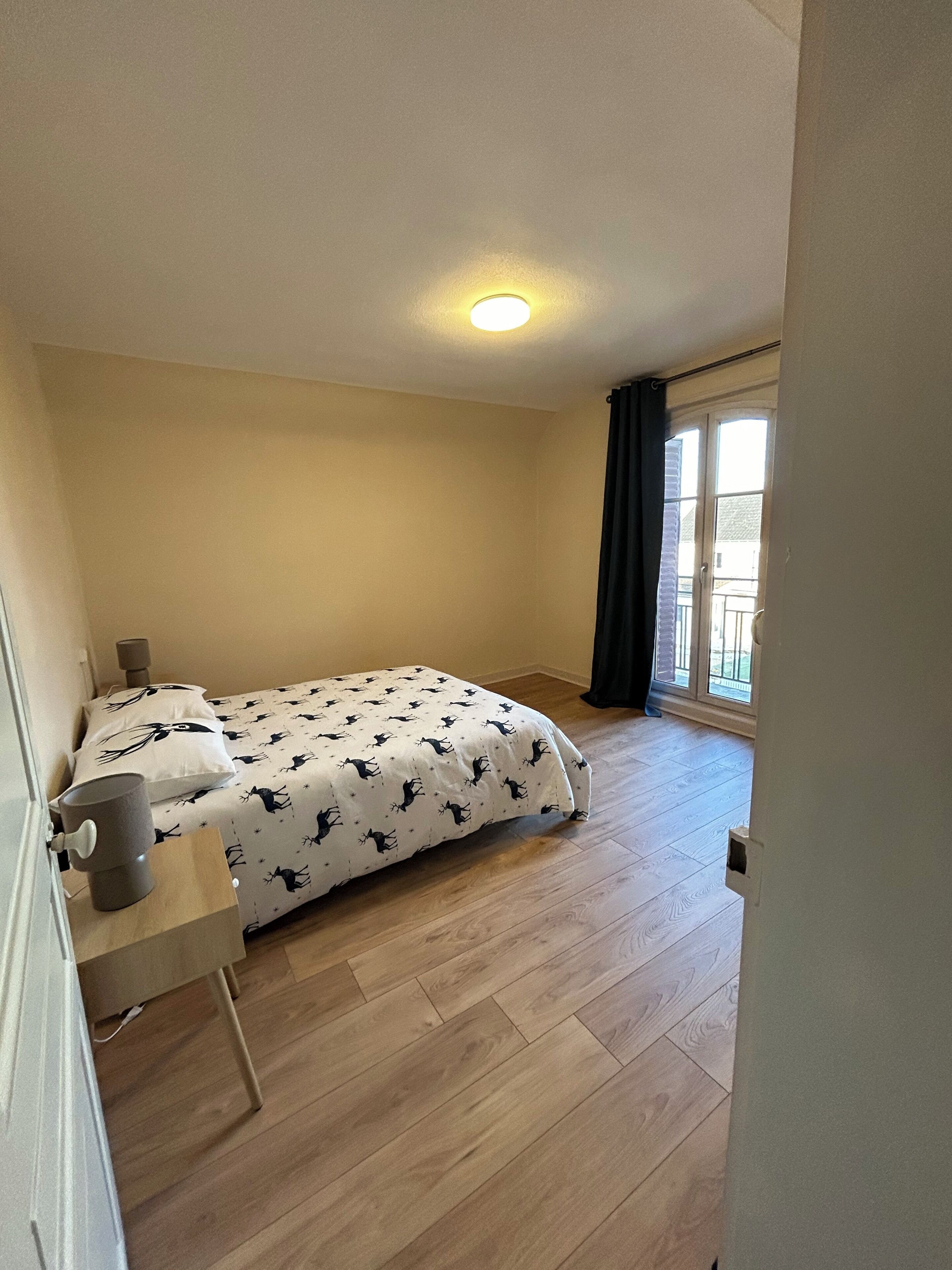 Une petite chambre à coucher, bien éclairée, avec un lit, une table de chevet et une fenêtre avec des rideaux.