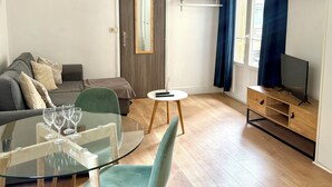 A modern living room with a glass dining table, teal chairs, a grey sofa, a TV on a wooden stand, and a large window with blue curtains. - Studio Carré d'Or with balcony (Nice)