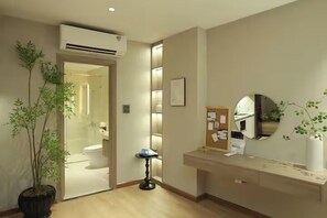 Ein Hotelzimmer mit Holzboden, einem Blumentopf, einem kleinen runden Tisch, einem Schminktisch mit Spiegel und einer Wandklimaanlage.