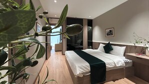 A modern bedroom with a large bed, a bedside table, and a nightstand. There is a plant in the foreground and a door leading to another room. - The Lumi Apartments by XOI Residences (Hanoi)