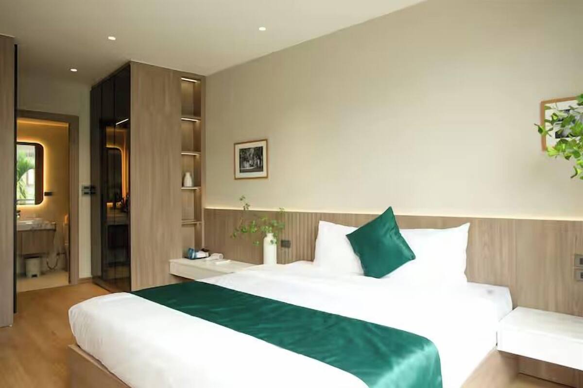 A modern hotel room with a large bed, bedside tables, a desk, and a bathroom visible through an open door.