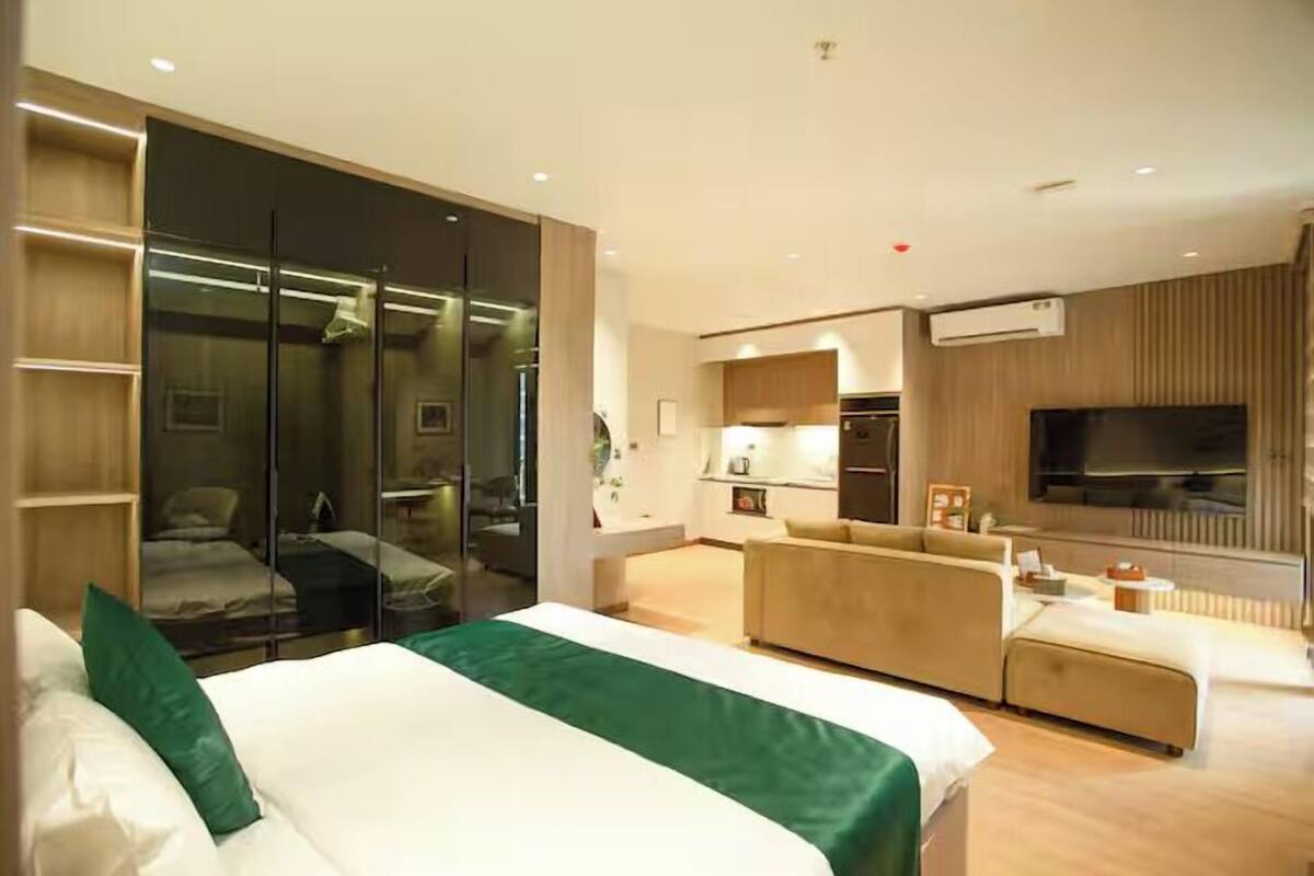 A modern hotel room with a large bed, a sofa, a TV, and a kitchen area.