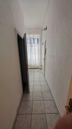 Ein schmaler Korridor mit Fliesenboden, weißen Wänden und einer Tür zu einem Raum mit Heizkörper und einem Fenster mit durchsichtigen Vorhängen.