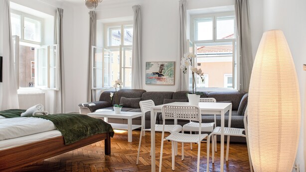 A modern hotel room with a bed, a sofa, a dining table, and chairs. There are windows with curtains, a chandelier, and a potted plant. - Group Stay Comfortable Multi Apart (Vienna)