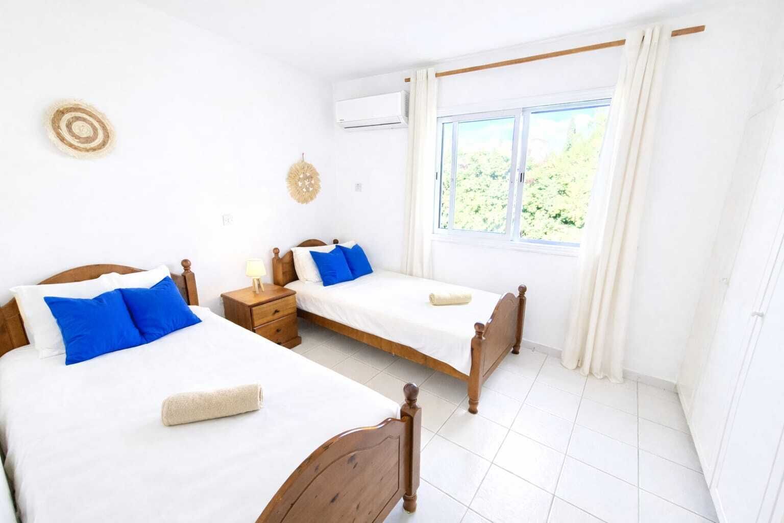 Une chambre propre et bien éclairée, avec deux lits en bois, du linge de lit blanc et une fenêtre avec des rideaux.