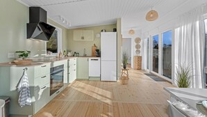 Ett modernt kök med vita skåp, en inbyggd ugn och ett kylskåp. Det finns en bänkskiva i trä med en diskho och ett fönster ovanför. Golvet är av trä, och det står en växt på fönsterbrädan.