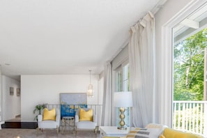 A bright living room with a white ceiling, a large window with white curtains, a painting on the wall, and a lamp on a side table.