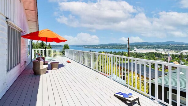 Eine Holzterrasse mit weißer Geländer, einem roten Sonnenschirm und Blick auf einen See und Berge.