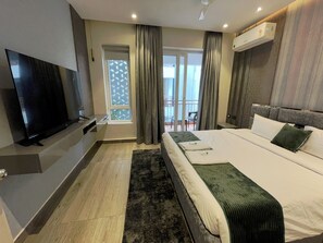 A modern hotel room with a large bed, a flat-screen TV, a balcony with a view, and an air conditioning unit. - Rohm Staycations (Gopalpur)