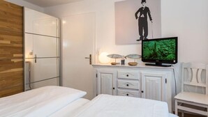 A hotel room with two beds, a television, a wooden wardrobe, and a white cabinet with decorative items. - Seerose Sylt - Modern 3-room apartment with dune view and balcony (Sylt)