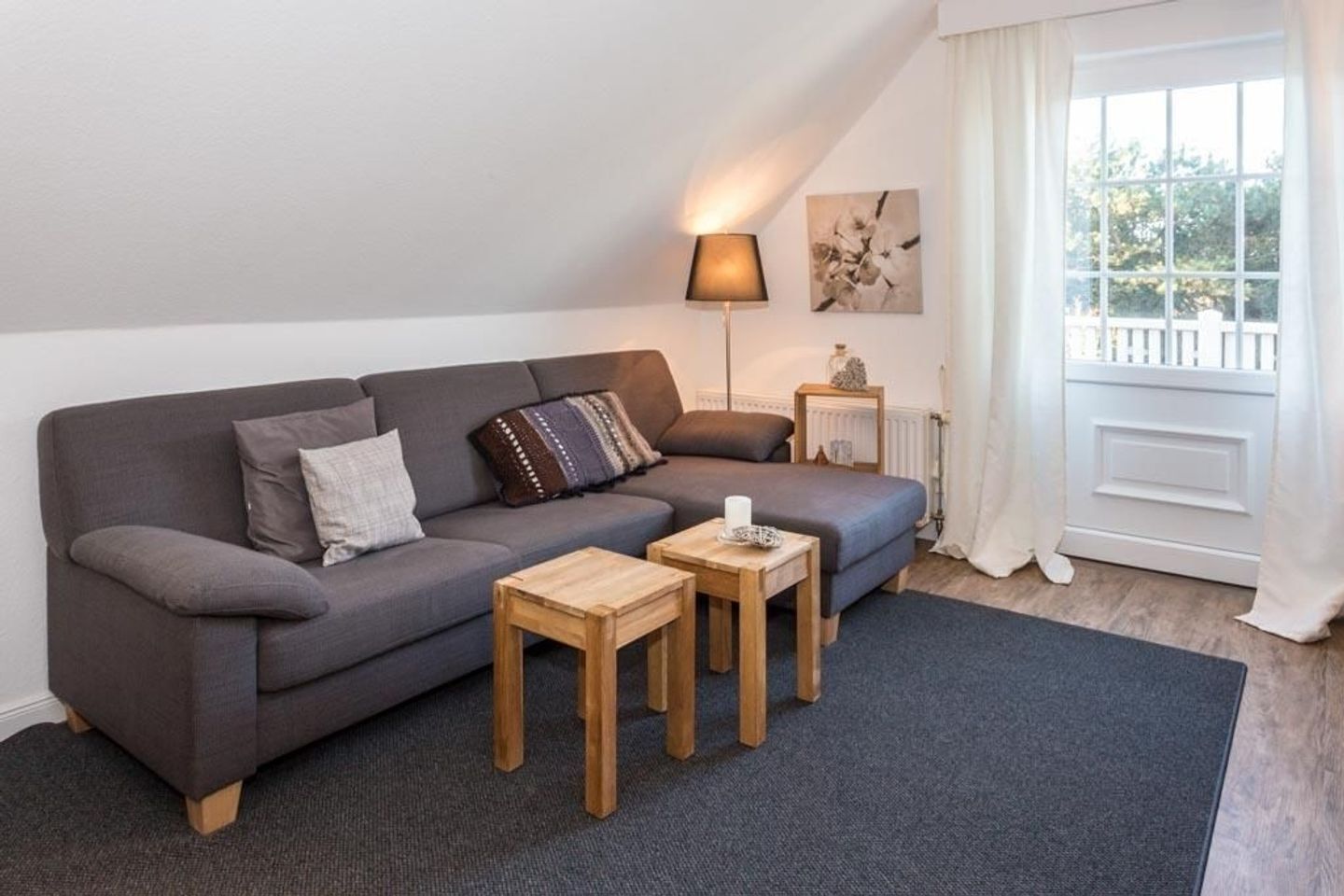 Un salon moderne avec un canapé gris, deux petites tables d’appoint en bois, une lampe sur pied et une fenêtre avec des rideaux.