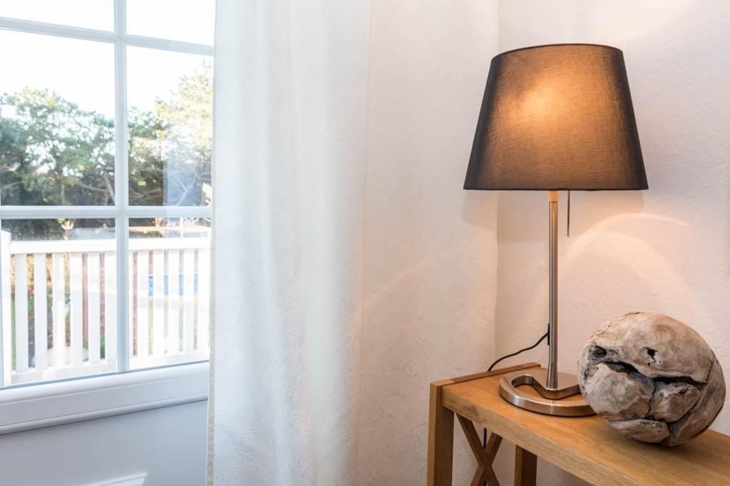 A lamp on a wooden table next to a window with a view of a white fence and trees.