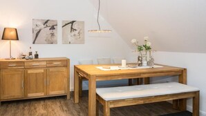 A dining area with a wooden table, bench, and a lamp. There are two framed pictures on the wall. - Seerose Sylt - Modern 3-room apartment with dune view and balcony (Sylt)