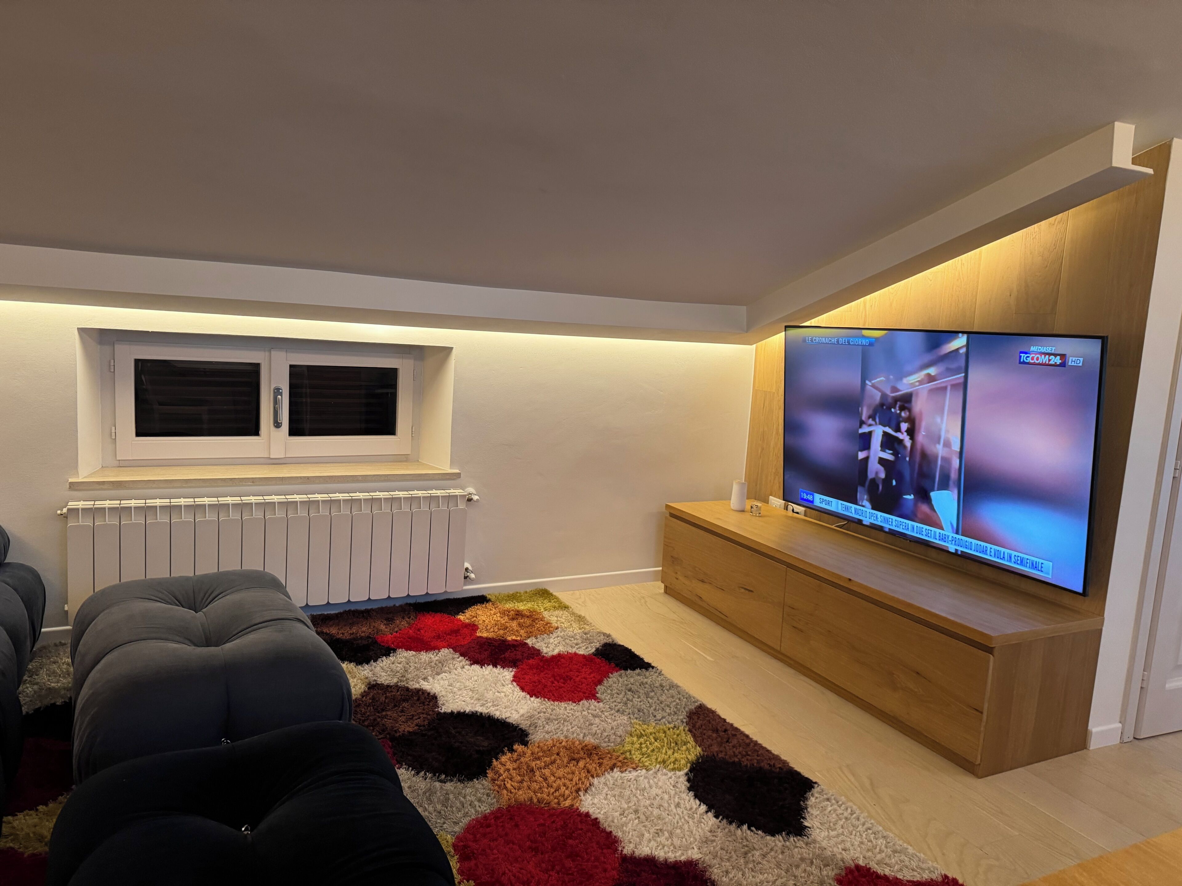 Una sala de estar moderna con un televisor de pantalla plana grande, una ventana con persianas, un radiador, un tapete estampado y un mueble de madera para entretenimiento.