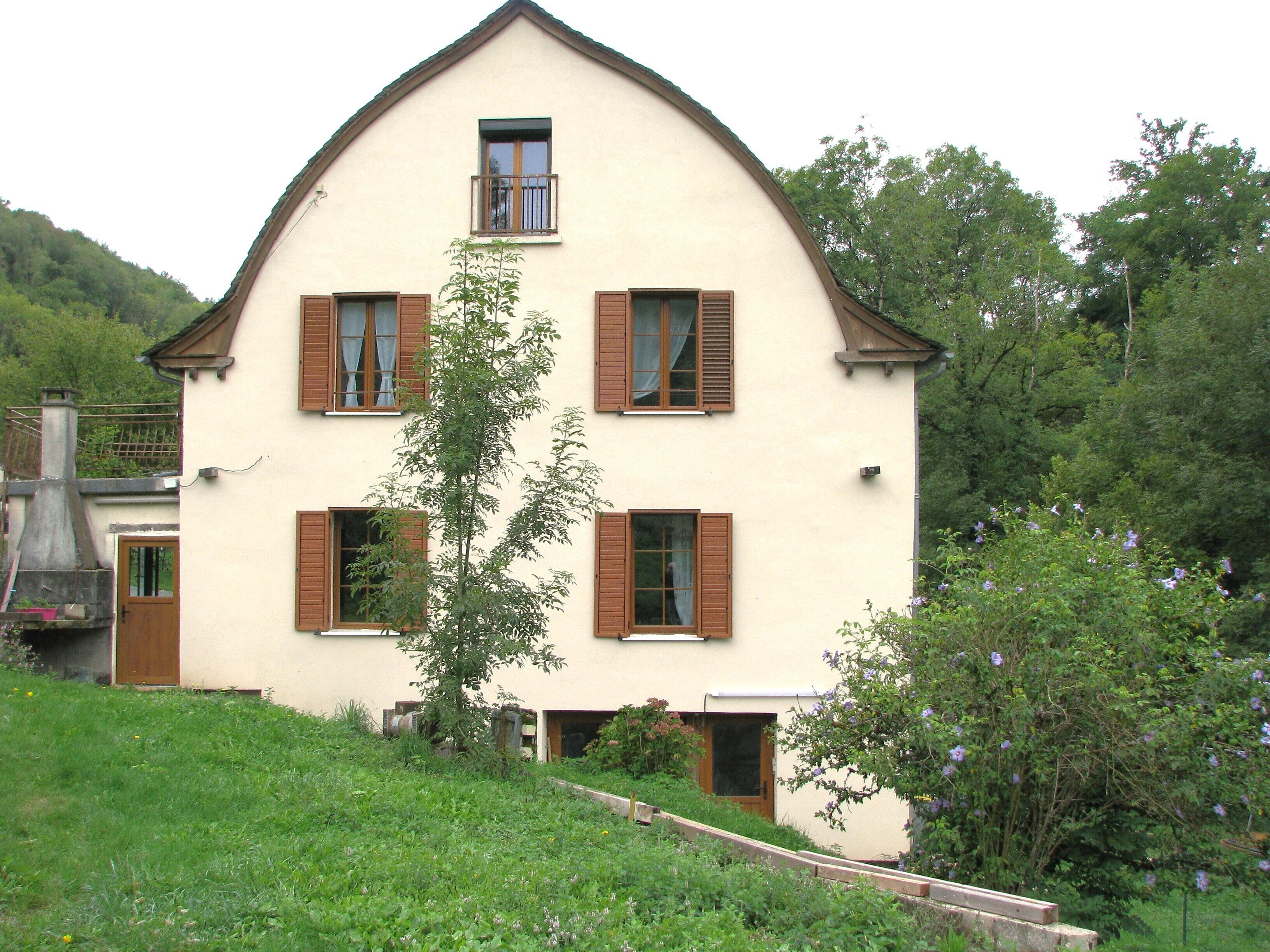 Une maison à deux étages, dotée d’un toit à pignon abrupt, de nombreuses fenêtres avec volets et d’un petit balcon.