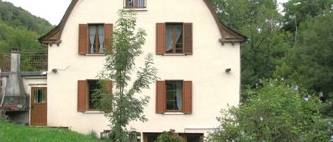 Une maison à deux étages, dotée d’un toit à pignon abrupt, de nombreuses fenêtres avec volets et d’un petit balcon.