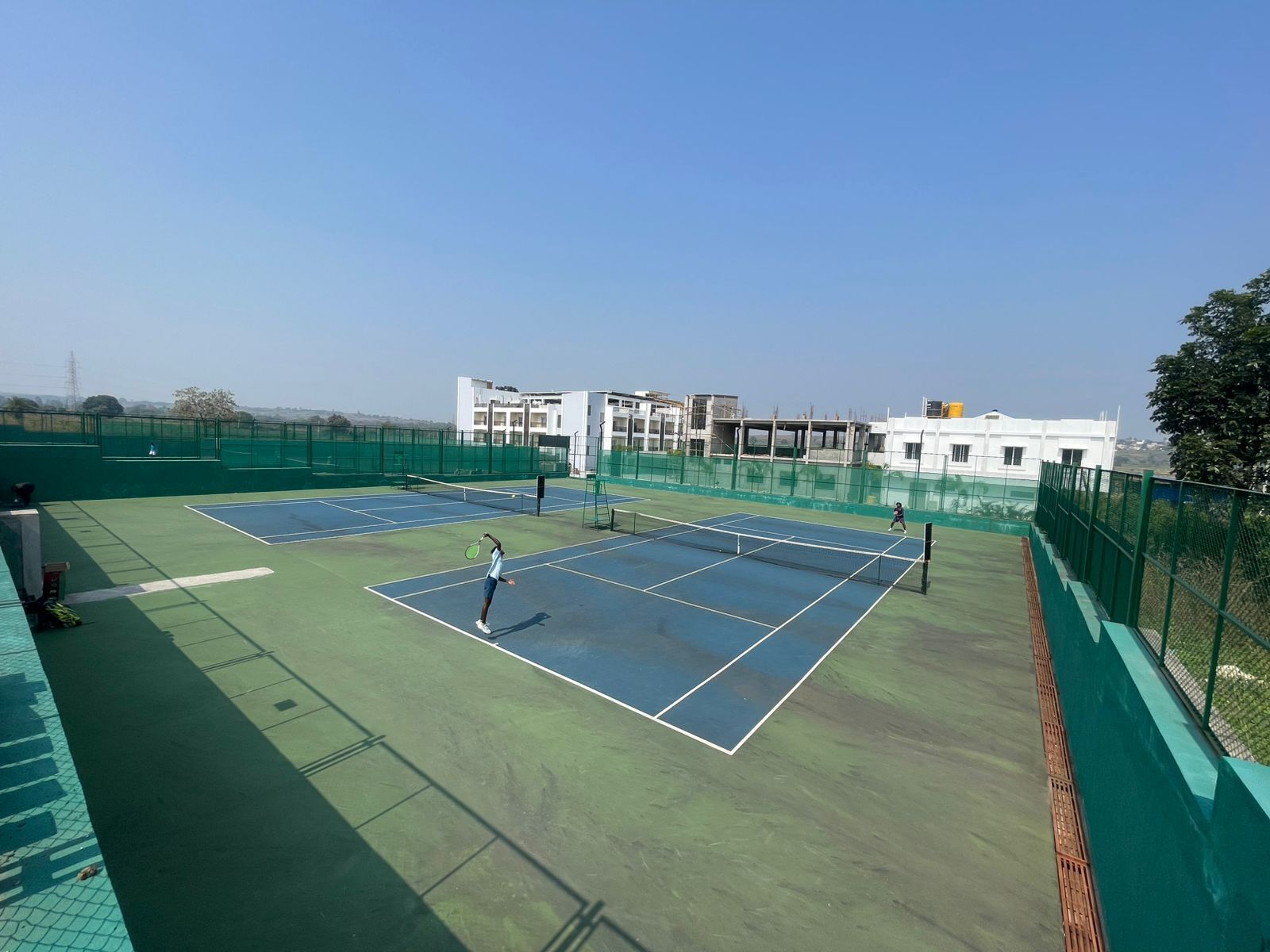 Un court de tennis comprenant deux terrains, entouré d’une clôture verte, avec des bâtiments en arrière-plan.