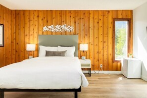 A modern bedroom with a wooden paneled wall, a large bed with white bedding, two bedside tables with lamps, and a window with a view of greenery. - Gateway Suites: Pet-friendly Getaway near Banff (Harvie Heights)