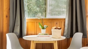 Une pièce avec une table ronde en bois, deux chaises blanches, une fenêtre avec des rideaux, une plante en pot et un sac de pain.