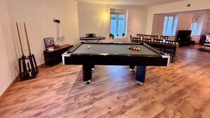 Une table de billard dans une pièce dont le sol est en bois, avec une fenêtre équipée de stores et un luminaire au plafond.