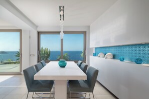 A modern dining area with a large table and chairs, a decorative vase, and a view of the ocean through large windows. - Caldera Blue Villa!! (Malevizi)