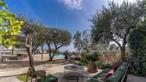 An outdoor patio with stone flooring, a round table, green cushioned seating, and a pool in the background. - Villa Chez Piè: Panoramic Views, Pool & Concierge (Massa Lubrense)